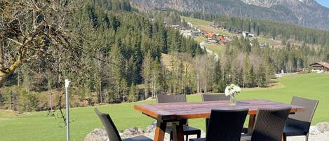 Un espace de restauration en plein air, avec une table et des chaises, situé devant un paysage de montagnes enneigées et d’une vallée boisée.