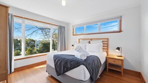 A bedroom with a large window, a bed with pillows and a blanket, a bedside table with a lamp, and a view of trees outside. - Coningham Beach House (Coningham)
