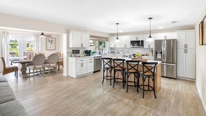 Una cocina amplia con armarios blancos, una isla central con taburetes de bar, un refrigerador y un comedor con mesa y sillas.