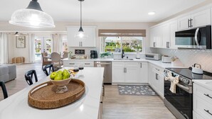 A modern kitchen with white cabinets, a central island with a woven tray, a dining area with a grey sofa, and a view of a garden through the window. - Brand New Saltwater Pool! - Waterfront Canal - Luxury Home (Apollo Beach)