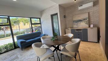 A modern living room with a blue sofa, a wooden dining table, and four chairs. There is a large window with a view of palm trees and a street.