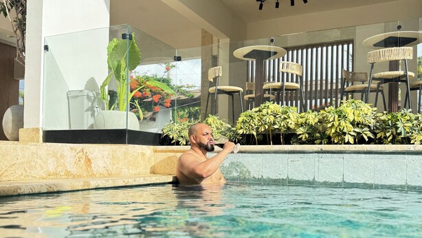 Un hombre en una piscina con cerramiento de vidrio, rodeado de plantas en macetas y mobiliario de exterior.