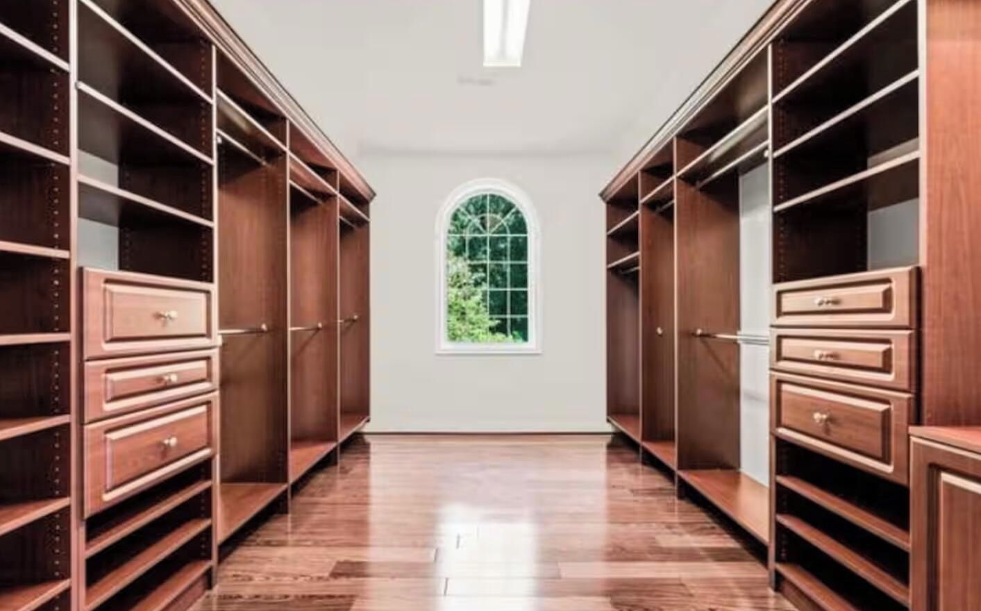 A walk-in closet with wooden shelving and drawers, a large arched window, and a polished wooden floor.