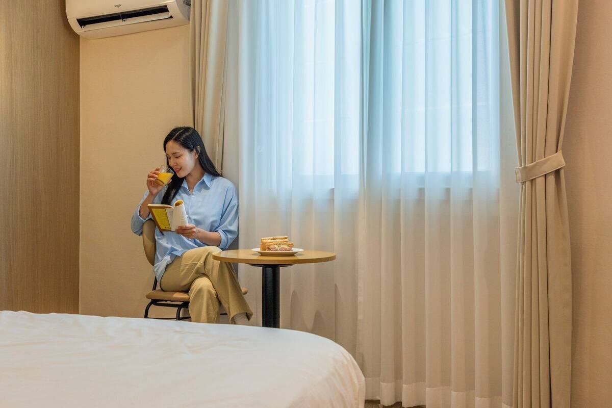 Ein Hotelzimmer mit einem Bett, einem kleinen Tisch mit einem Sandwich und einer Person, die ein Buch liest.