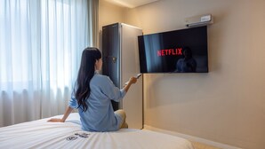 A person sitting on a bed in a hotel room, holding a remote control, with a television displaying the Netflix logo.