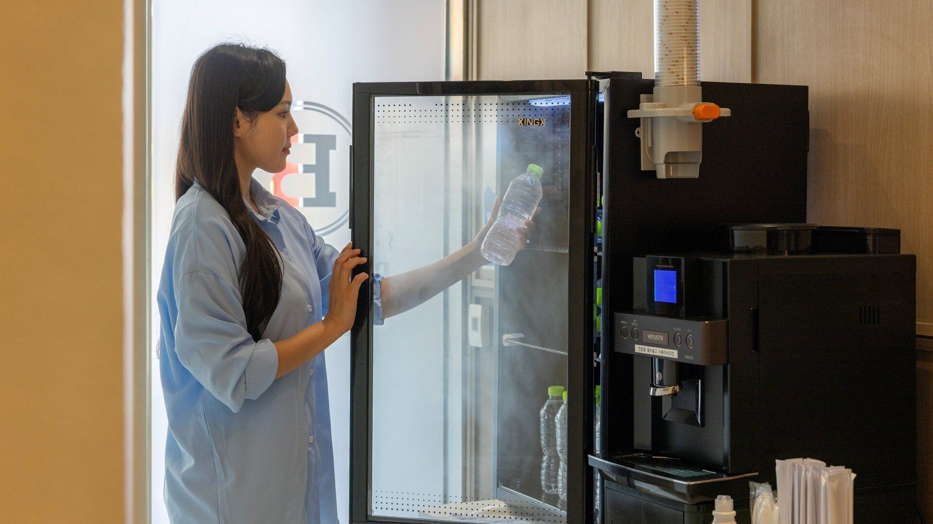 A person interacting with a water dispenser in a room with a glass door and a control panel.