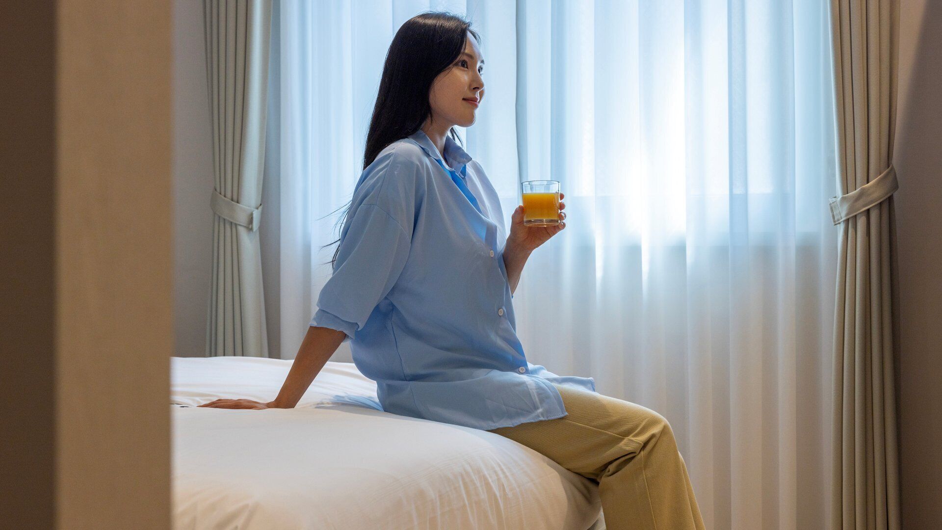 A person sitting on a bed in a room with sheer curtains and a window.