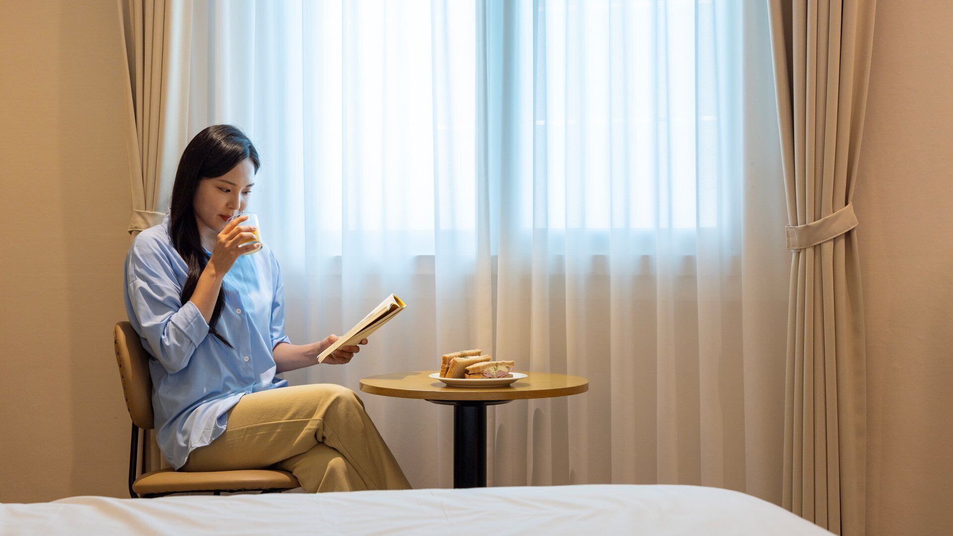 A person sitting on a bed in a room with a window, holding a book and a glass of drink, with a small table beside them.