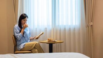 A person sitting on a bed in a room with a window, holding a book and a glass of drink, with a small table beside them.
