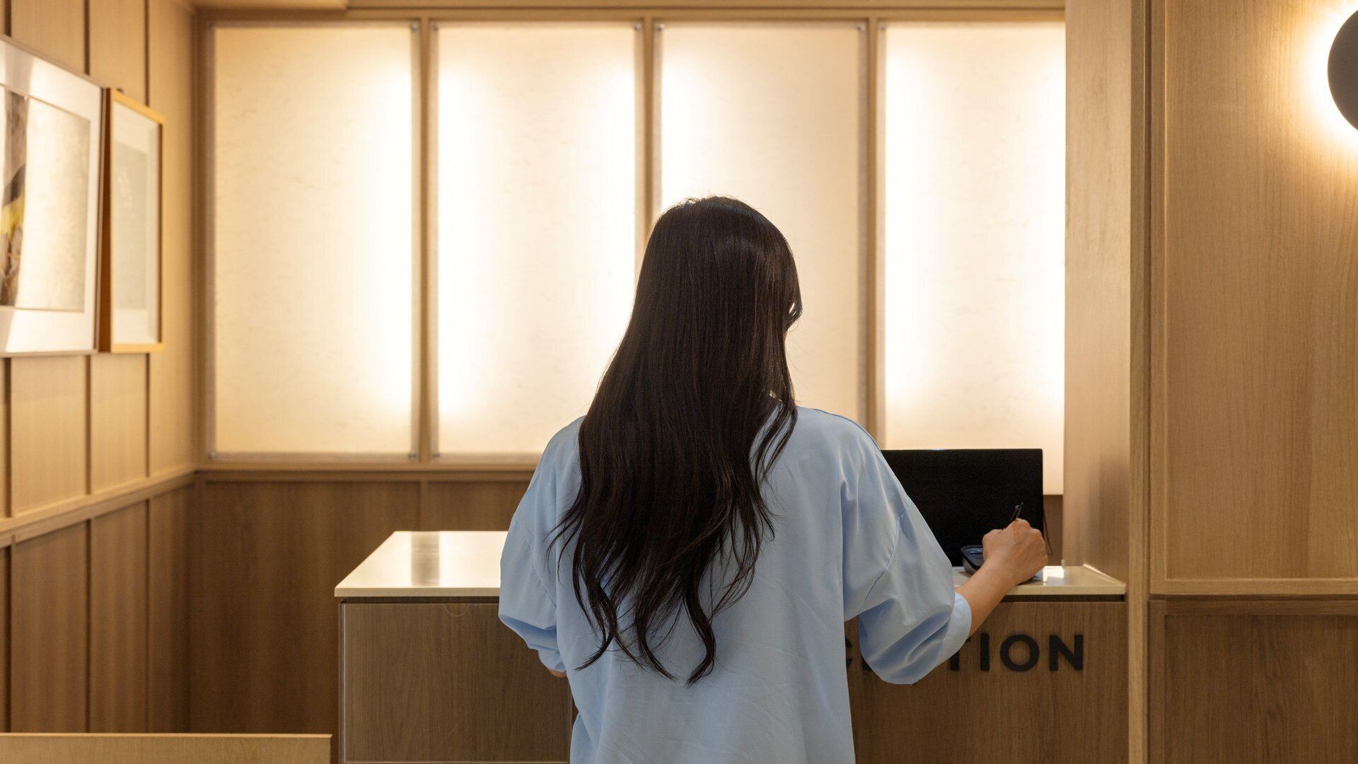 Una persona de pie frente a la recepción en el vestíbulo de un hotel con paneles de madera y una gran pared iluminada.