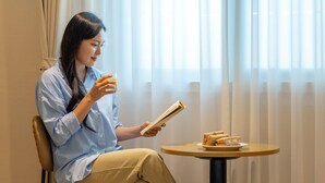 A person sitting by a window with sheer curtains, holding a book and a glass of orange juice, with a plate of sandwiches on a small round table.