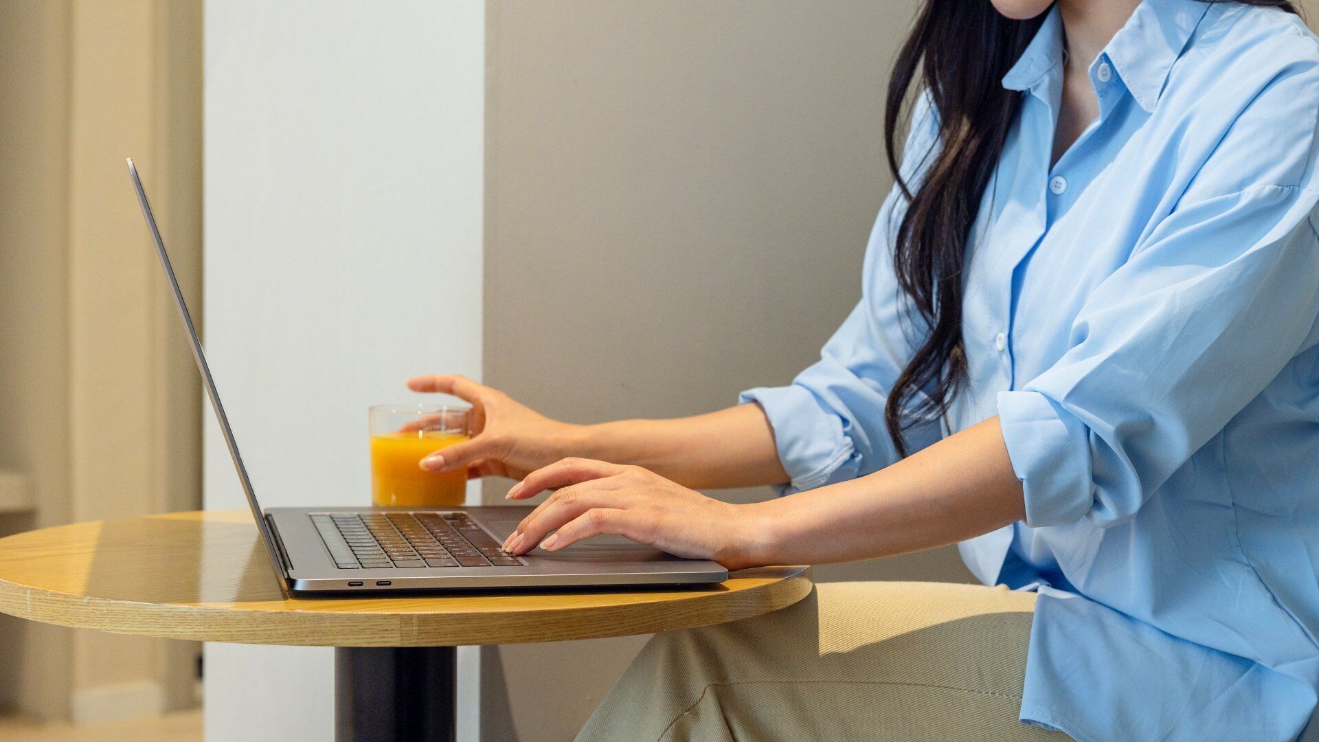 A person using a laptop on a round wooden table with a glass of orange juice.