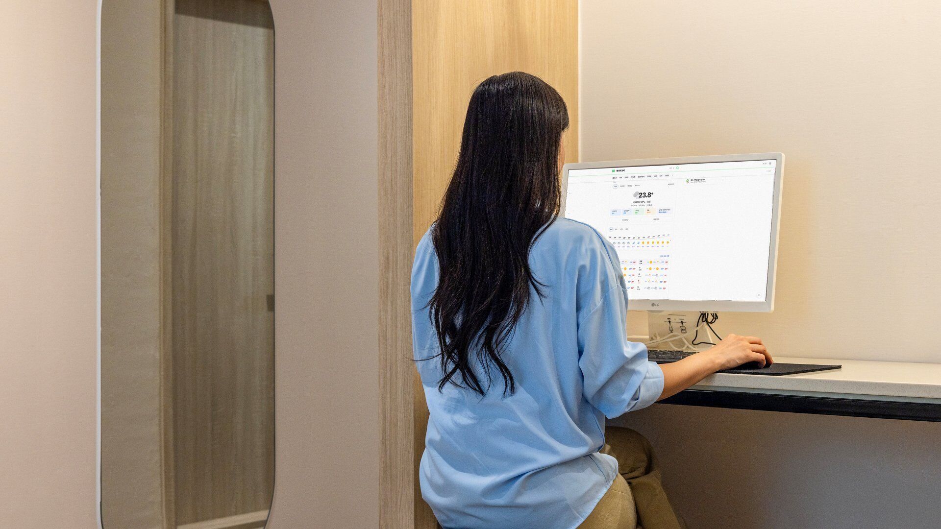 Une personne assise à un bureau devant un ordinateur, dans une pièce dotée d’une porte en bois et d’un mur de couleur claire.