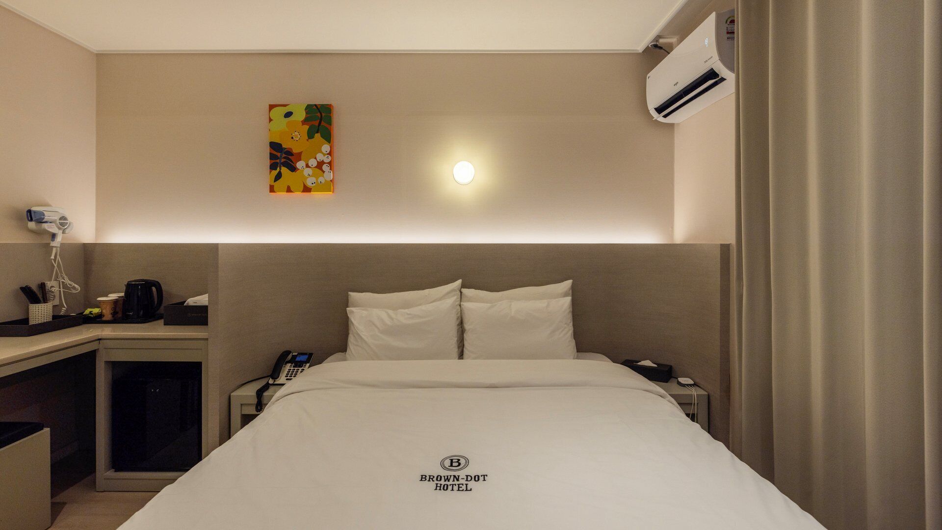 A hotel room with a bed, bedside table, a phone, an air conditioning unit, and a painting on the wall.