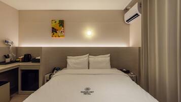 A hotel room with a bed, bedside table, a phone, an air conditioning unit, and a painting on the wall.