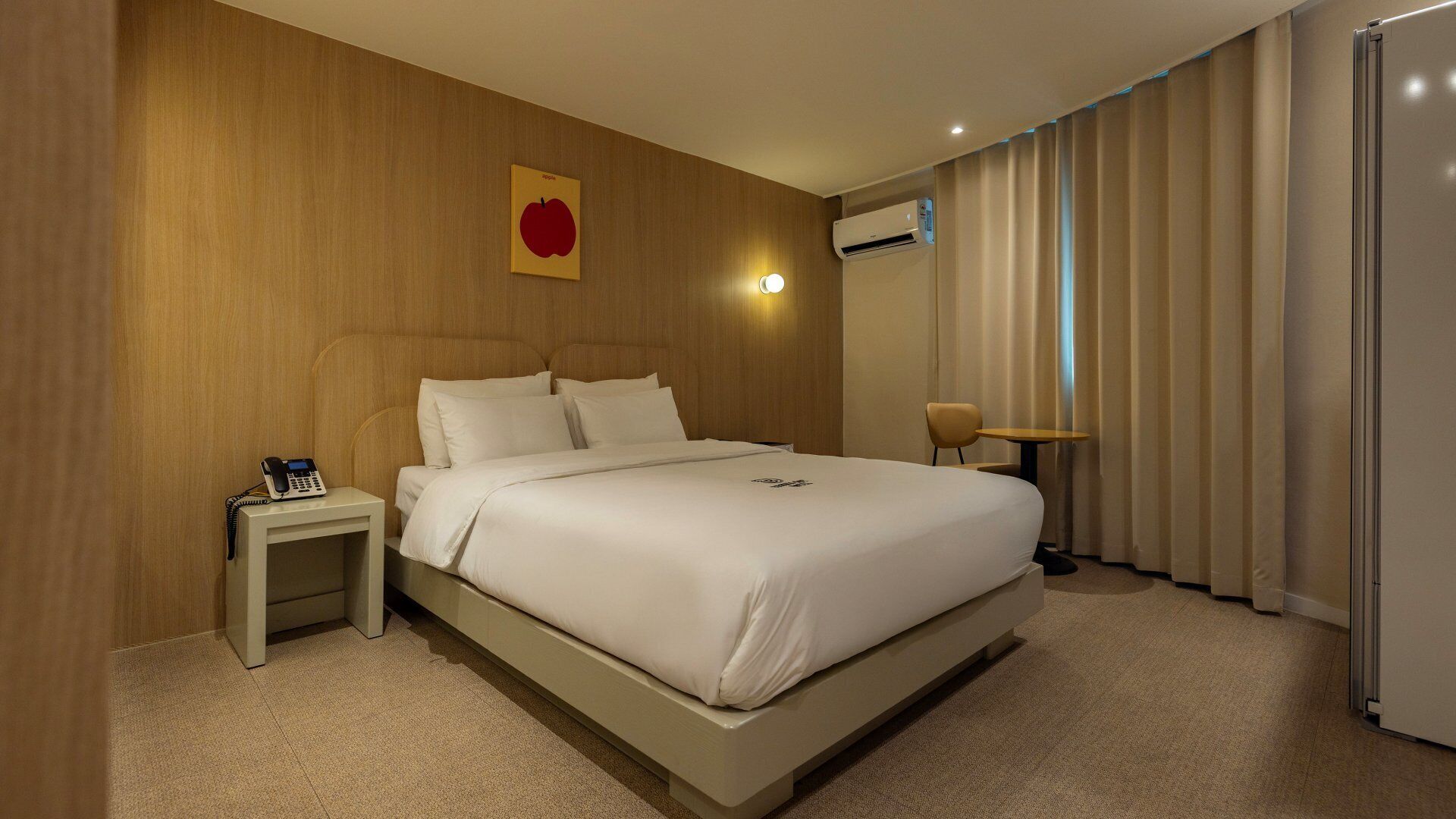 A hotel room with a large bed, a bedside table with a phone, a wall-mounted air conditioner, and a window with curtains.