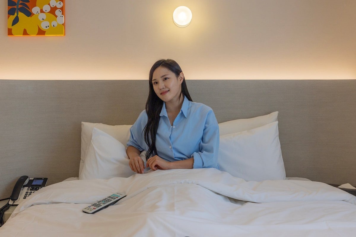 Ein Hotelzimmer mit Bett, weißer Bettwäsche, einer Fernbedienung, einem Nachttischtelefon und einer Wandleuchte.