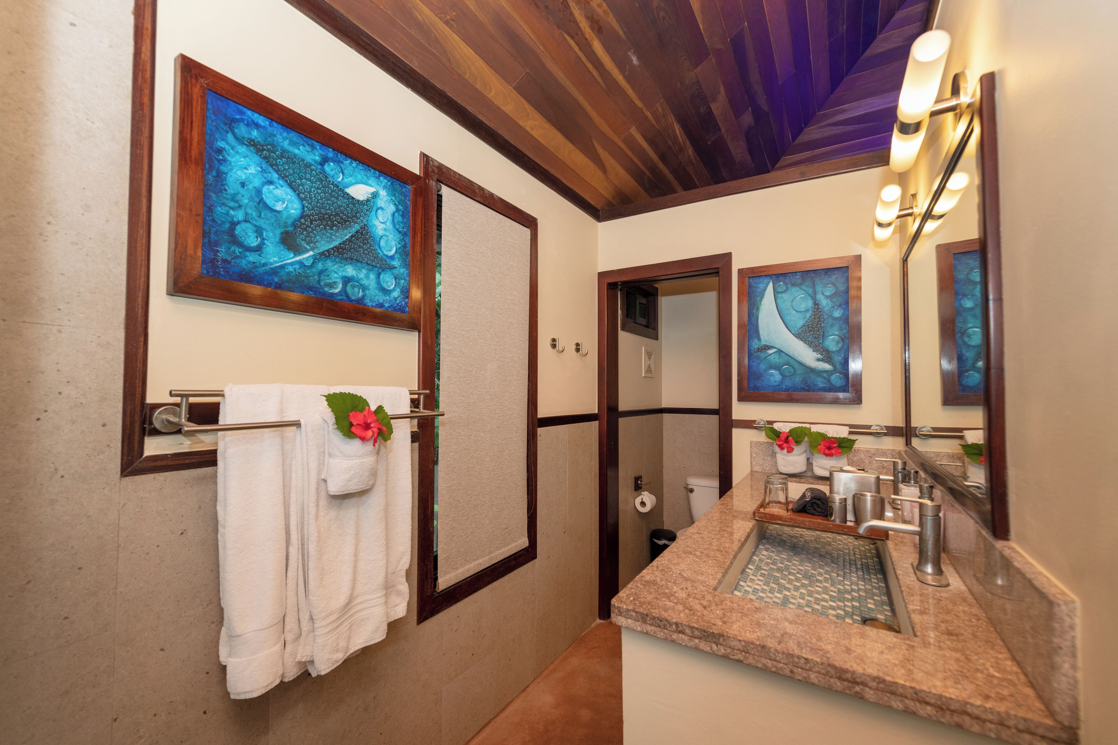 Une salle de bain avec un plafond en bois, un lavabo doté d’un plan de travail en granit et deux œuvres d’art encadrées au-dessus du miroir.