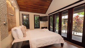 A bedroom with a large bed, wooden ceiling, a TV mounted on the wall, and a view of the outdoors through large windows.