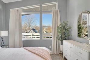 A bedroom with a large window, a bed, a dresser, and a potted plant.