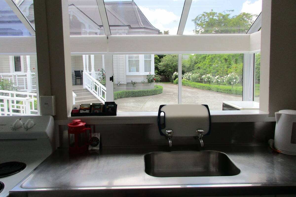 A kitchen with a stainless steel sink, a coffee maker, and a window overlooking a garden and a house.