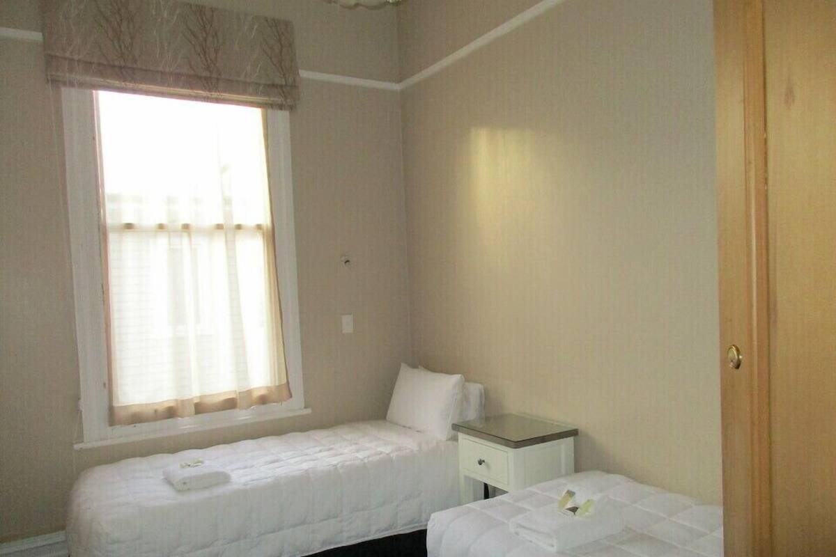 A small, clean room with two beds, a window with curtains, a bedside table, and a wooden door.