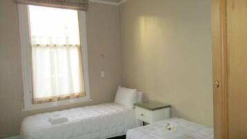 Una habitación pequeña y limpia con dos camas, una ventana con cortinas, una mesita de noche y una puerta de madera.