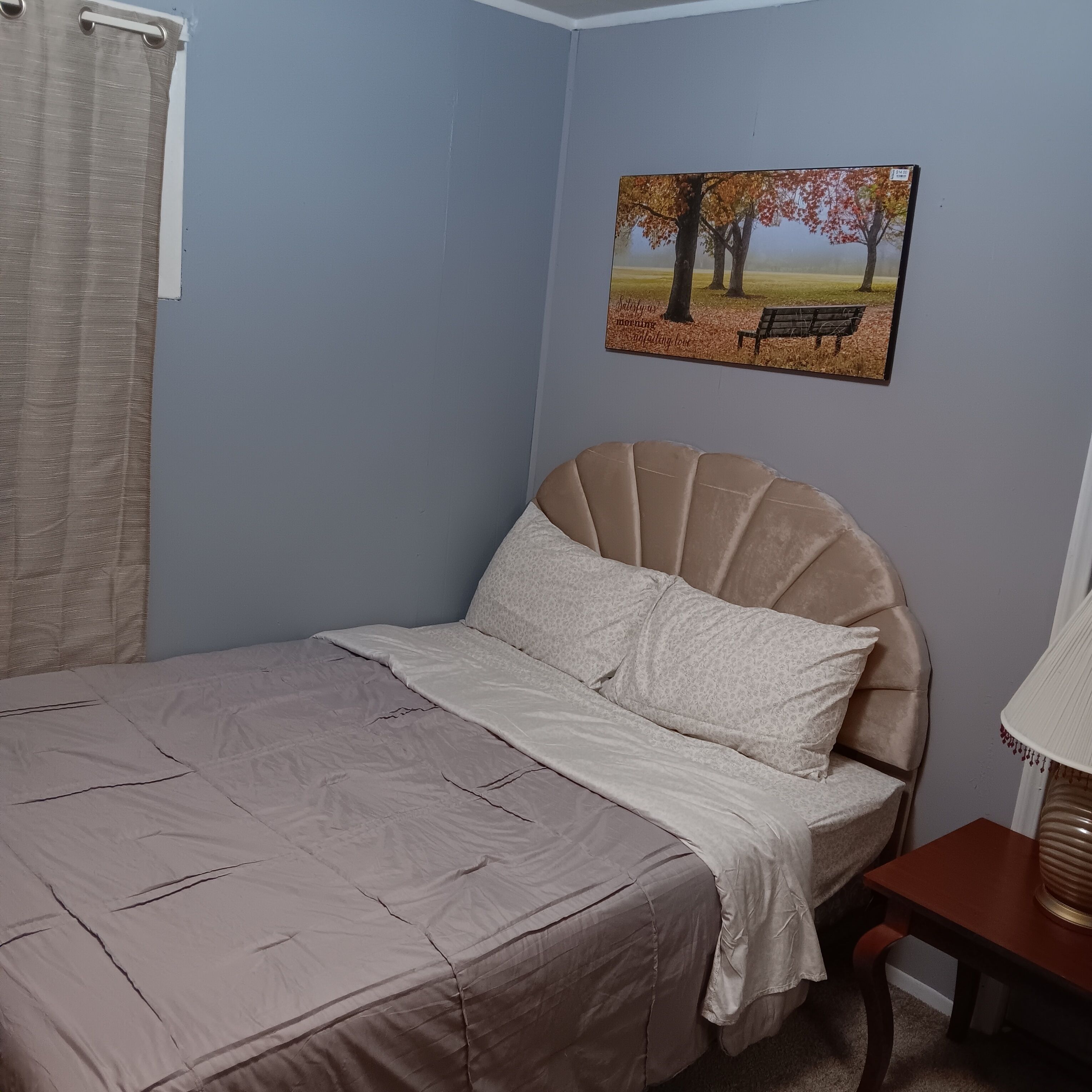 A bedroom with a bed, a painting of trees, and a lamp.