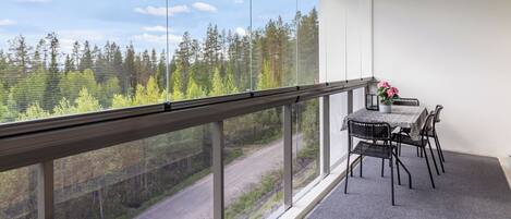 A balcony with a table and chairs, overlooking a forested area with a road.