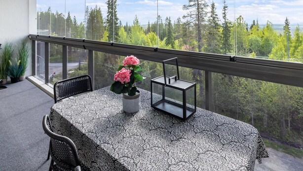 Un balcón con una mesa y sillas, un florero con flores y vista a un bosque.