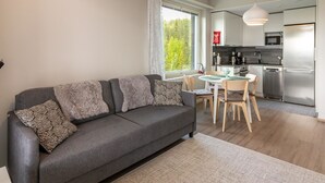 Una sala de estar moderna con un sofá gris, cojines estampados, una mesa de comedor con sillas y una zona de cocina con electrodomésticos de acero inoxidable.