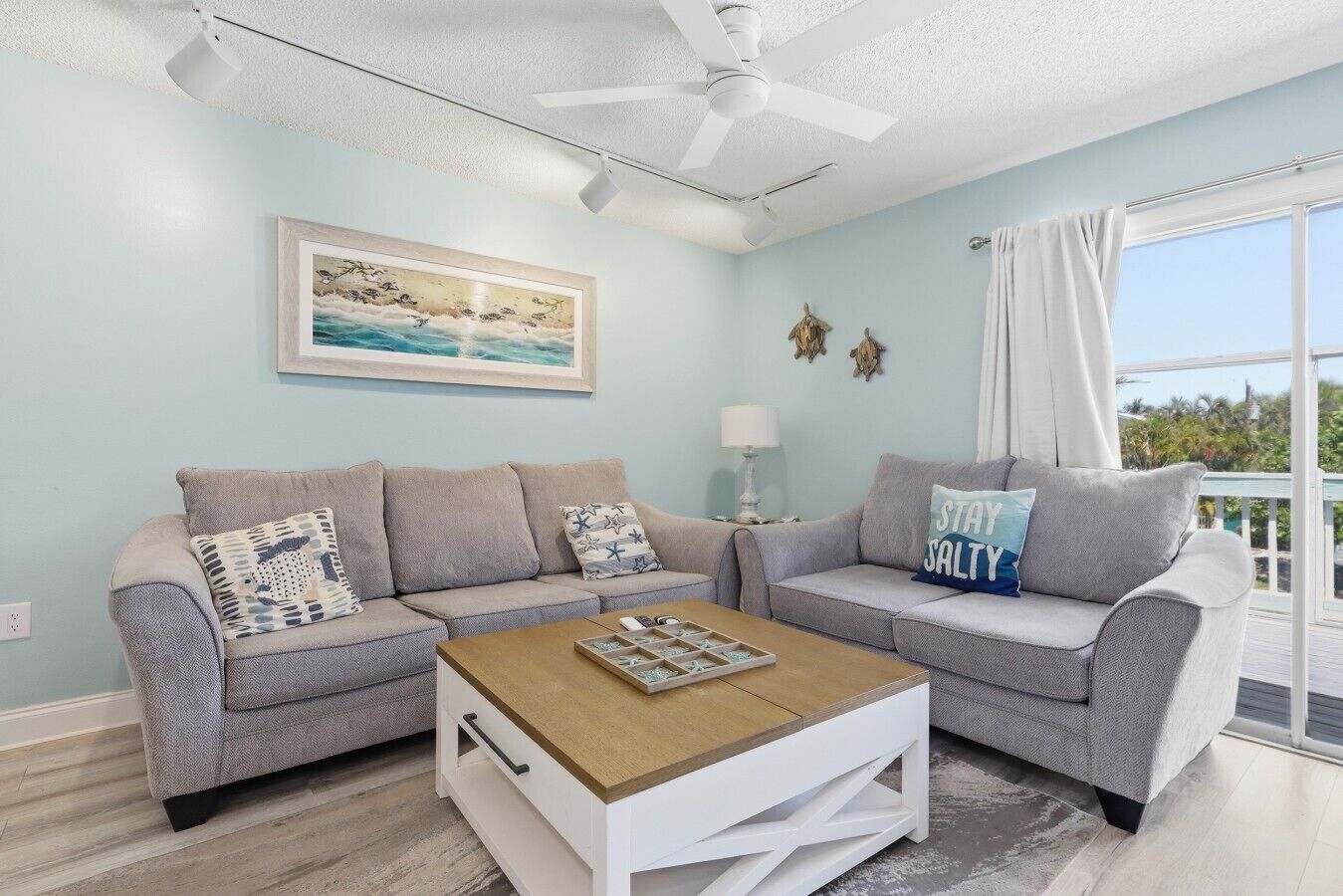 A living room with two grey sofas, a wooden coffee table, a ceiling fan, a framed picture, and a view through a window.
