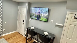A room with a dining table, two chairs, a television mounted on the wall, and a patterned rug on the floor. - Rental studio unit in Queen Village of Philadelphia (Philadelphia)