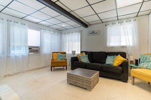 A living room with a sofa, armchair, and ottoman, large windows with curtains, and a ceiling with a grid pattern. - The Pietroman: Cottage in Sunset Bay with Fire Pit (Sunset Bay)