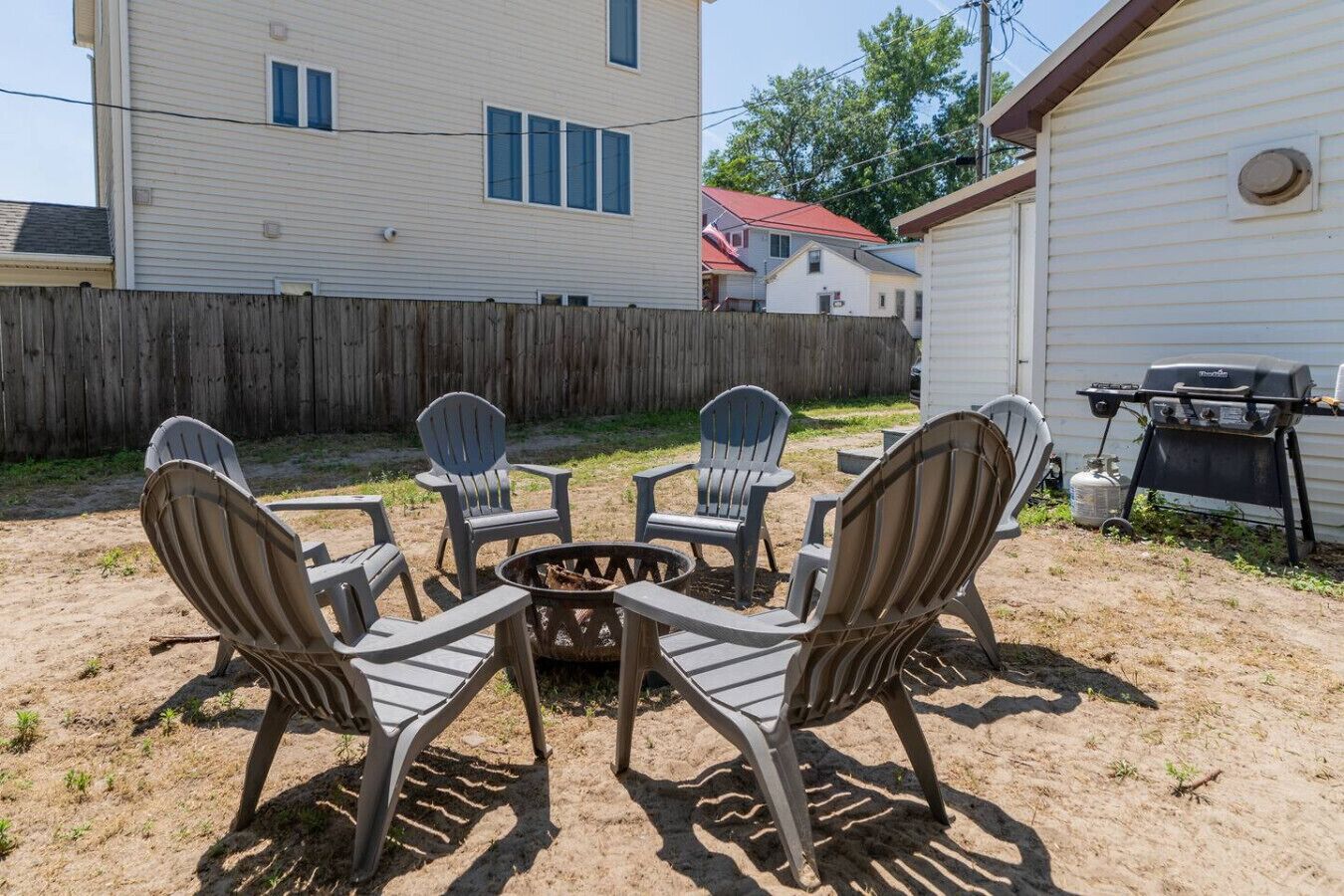 A backyard with a fire pit surrounded by four Adirondack chairs, a barbecue grill, and a wooden fence.