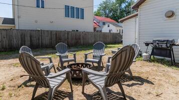 A backyard with a fire pit surrounded by four Adirondack chairs, a barbecue grill, and a wooden fence.