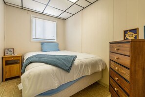 A bedroom with a bed, a nightstand, a dresser, a window with blinds, and a ceiling with a grid pattern. - The Pietroman: Cottage in Sunset Bay with Fire Pit (Sunset Bay)