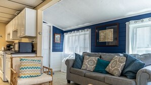 A cozy living room with a grey sofa, a wooden chair, and a small table. There is a kitchen area with white cabinets and a microwave. - Remi-Loo's Bay Escape: Cottage in Sunset Bay (Sunset Bay)