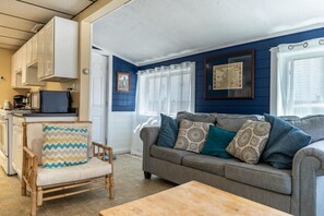 A cozy living room with a grey sofa, a wooden chair, and a small table. There is a kitchen area with white cabinets and a microwave. - Remi-Loo's Bay Escape: Cottage in Sunset Bay (Sunset Bay)