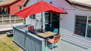 Een overdekt terras met een tafel, stoelen en een rode parasol, met uitzicht op een omliggend huis.