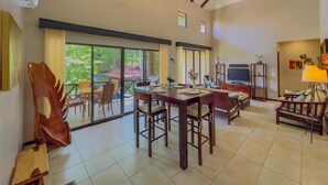 A spacious living area with a dining table, chairs, a sofa, a TV, and a large window offering a view of greenery. - Sombras #704 Beachside Escape Spacious 3BR Condo with Terrace (Playas del Coco)
