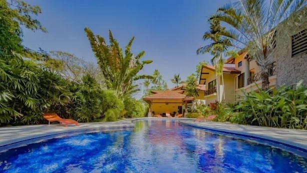 A swimming pool with a clear blue water surface, surrounded by lush greenery and a building with a red-tiled roof. - Sombras #704 Beachside Escape Spacious 3BR Condo with Terrace (Playas del Coco)