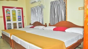 Un letto matrimoniale con struttura in legno, due letti singoli con struttura in legno, un cuscino rosso e bianco, una coperta gialla, una coperta bianca, una tenda grigia, un condizionatore d'aria bianco e una bacheca con manifesti motivazionali.