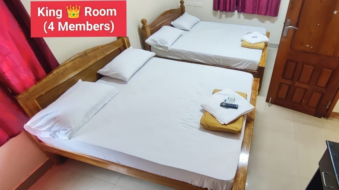 A King Room with two single beds, each with a wooden frame and white bedding. There is a small table with a yellow cloth and a black object on it. The room has a wooden door and a pink curtain.