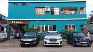 Un edificio a due piani con un cartello che indica 'JR Residency', segnalando che si tratta di una proprietà residenziale. Sono parcheggiate tre auto davanti e l'edificio ha diverse finestre e un condizionatore d'aria sulla parete esterna.