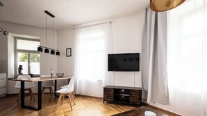 A modern living room with a wooden dining table, white chairs, a TV on a wooden stand, and a kitchen in the background. - FirstClass 2-room apartment (Leipzig)