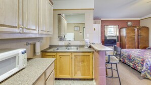 Una cocina compacta con microondas, pileta y armarios de madera. Hay un pequeño comedor con una silla y una cama con colcha estampada.