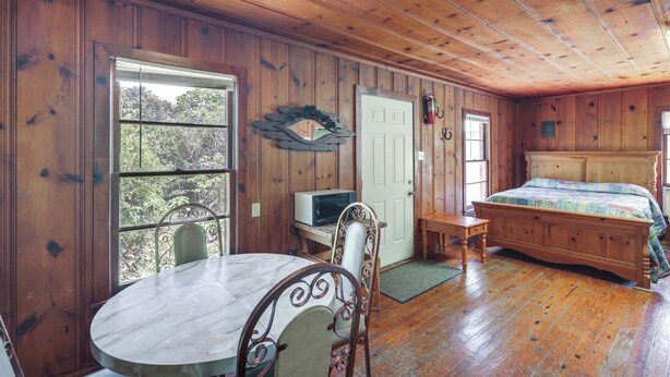 A wooden-paneled room with a bed, a dining table with chairs, a small desk, and a television. - On-Site Trails! Cozy Whitney Cottage 2 Mi to Lake (Whitney)