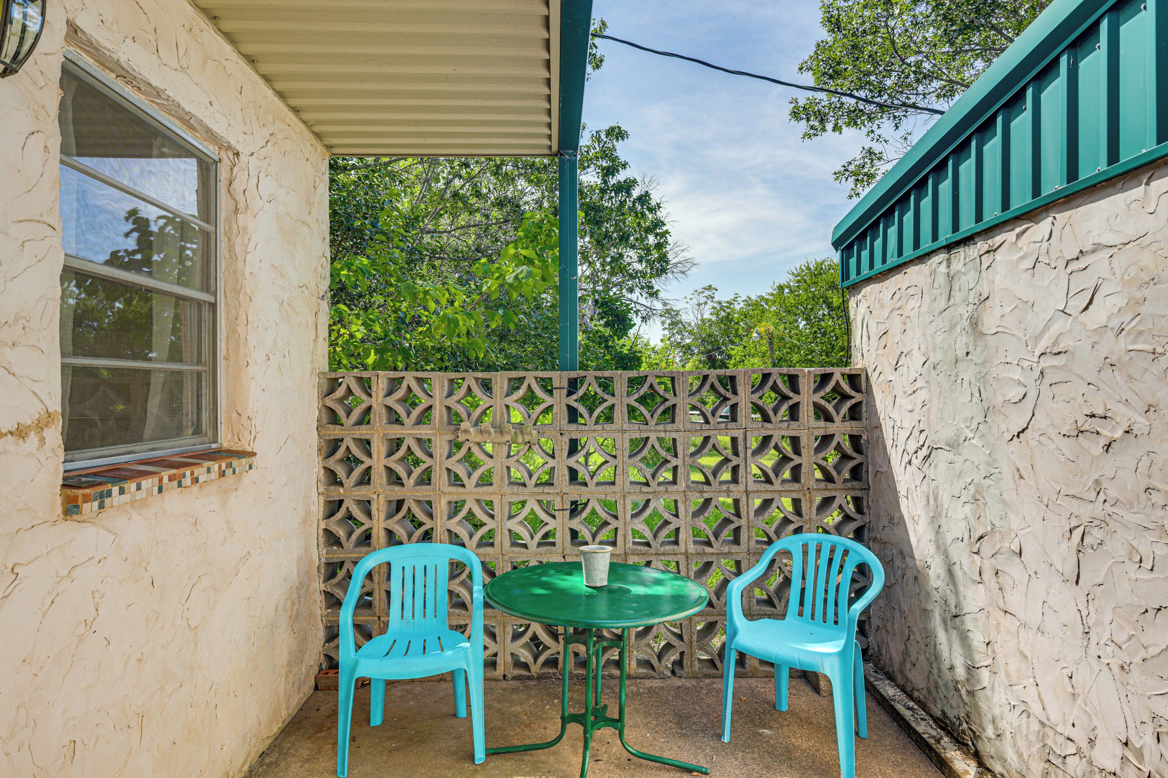 Un pequeño patio con dos sillas de plástico turquesa y una mesa verde, rodeado por un muro de piedra y un cerramiento ajardinado.