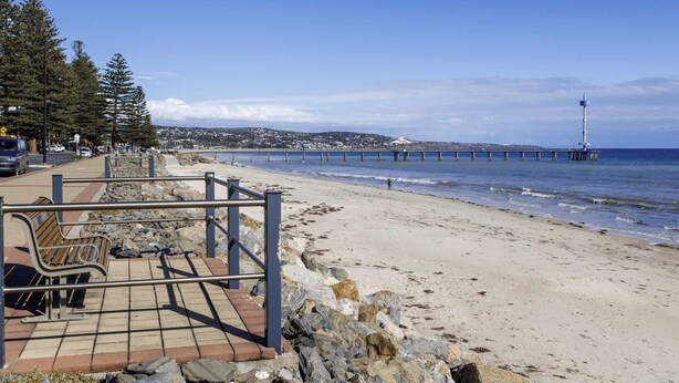 A beach with a pier extending into the water, a rocky shoreline, and a paved walkway with benches.
